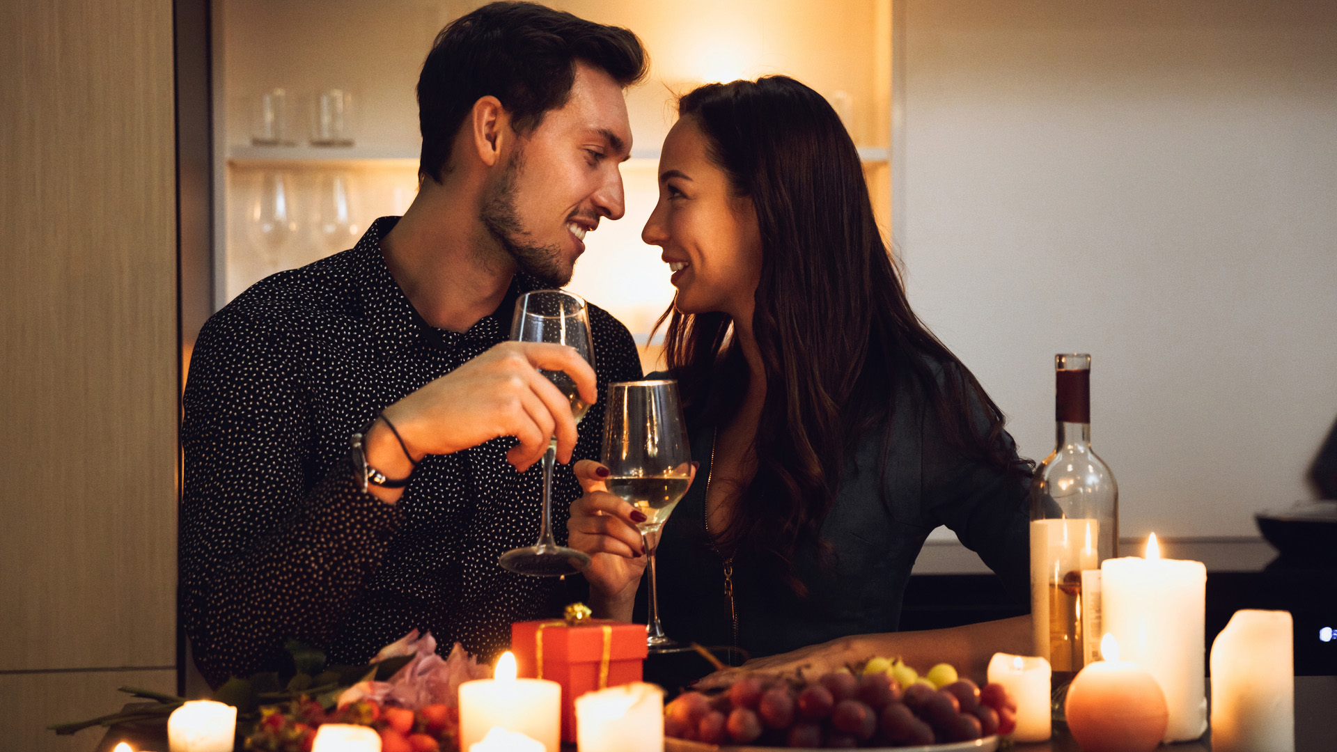 Brunette man and woman sitting close to each other while holding wine glasses and looking into each others' eyes. Lit candles